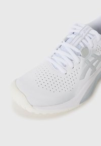 Hvid atletiksko med et mesh-overdel med ventilationshuller, grå accenter og en struktureret sål for greb. Snørebåndene er hvide og flade.