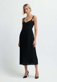 Frau mit kurzen blonden Haaren, die ein schwarzes, plissiertes Midikleid und schwarze High Heels trägt, steht vor einem schlichten weißen Hintergrund.