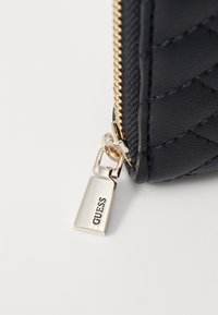 Portefeuille noir matelassé avec une fermeture éclair dorée et une étiquette suspendue portant le logo "GUESS". Le matériau semble synthétique avec une texture lisse.