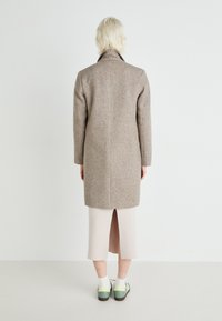 Hnedý kabát z vlnenej zmesi s vysokým golierom, rovný strih a dlhé rukávy, skombinovaný s svetlosivou sukňou a topánkami s zelenými akcentmi.