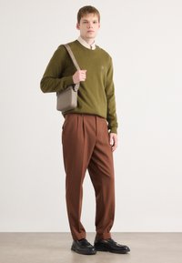 Olivgrüner Pullover, braune plissierte Hose und schwarze Schuhe. Trägt eine kleine beigefarbene Umhängetasche. Neutrale Hintergrund.