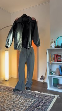 Person som håller upp en svart skinnjacka på en galge, klädd i grå byxor och svarta skor, stående på en mönstrad matta i ett vardagsrum.