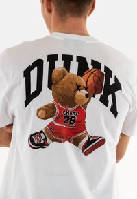 Camiseta blanca con una impresión gráfica de un oso pardo en una camiseta de baloncesto roja, sosteniendo un balón de baloncesto. Texto en negrita negro "DUNK" arriba.