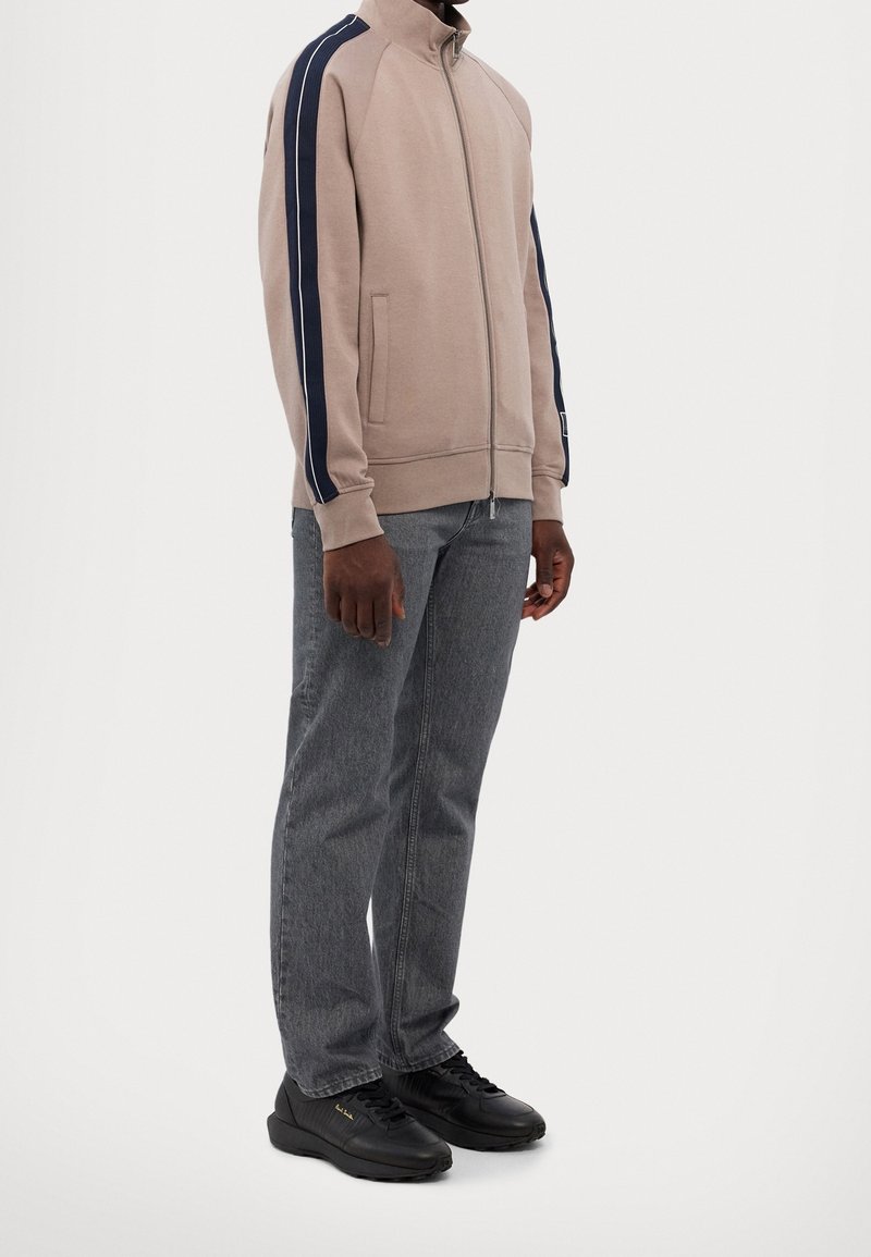 Homme portant une veste beige zippée avec des bandes bleu marine sur les manches, un jean droit gris et des baskets noires, debout sur fond blanc.