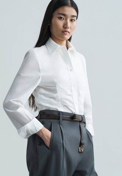 Bijela košulja na dugme s čvrstim kragnom i skupljanom teksturom, u kombinaciji s sivo prilagođenim hlačama koje imaju remen i bočni gumb.