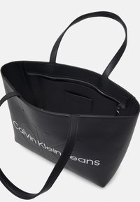 Melnā mākslīgās ādas tote soma ar iegravētu logotipu, dubultām siksnām un plašu iekšpusi, kurā ir rāvējslēdzēja kabata un strukturēta oderē.