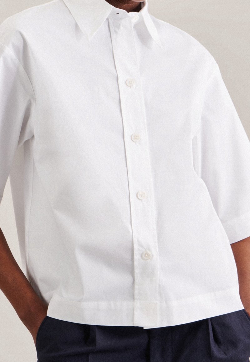 Vit kortärmad knäppskjorta med klassisk krage, slät textur och rak passform, med sex knappar framtill.