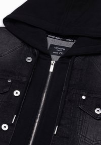 Veste noire à capuche avec fermeture éclair argentée, boutons pression en métal, poches avant et une étiquette taille M à l’intérieur du col.
