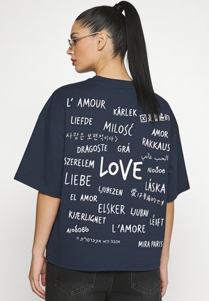Woman with glasses and ponytail wears a navy shirt with the word "love" written in multiple languages on the back.