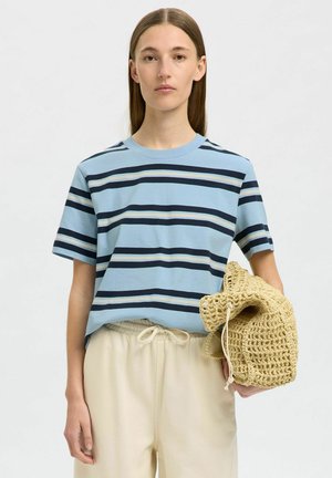 T-shirt bleu à rayures à manches courtes avec des rayures marine, crème et beige. Le mannequin tient un sac en crochet beige et porte un pantalon clair.