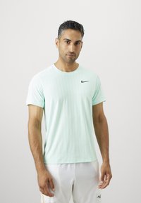 Mees lühikeste tumedate juustega, kes kannab helesinist Nike spordit-särk ja valgeid sportlikke lühikesi pükse, seisab lihtsa valge tausta ees.
