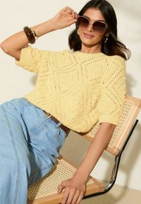 Jersey de punto amarillo con patrón texturizado, mangas cortas y corte holgado, combinado con vaqueros de mezclilla azul claro. El modelo también lleva gafas de sol de gran tamaño.