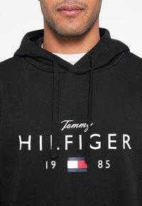Zwarte hoodie met verstelbare trekkoorden. Voorzien van witte tekst "Tommy HILFIGER" en een klein logo met rood-wit-blauwe strepen.