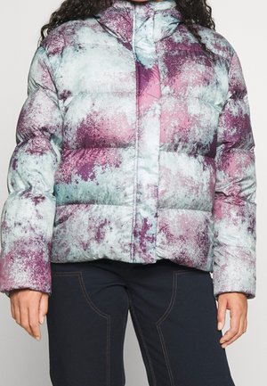Gros plan d'une personne portant une veste matelassée à capuche avec un motif abstrait violet et bleu clair et un pantalon noir.
