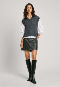 Gilet grigio lavorato a maglia con motivo a diamante sopra una camicia bianca, abbinato a una gonna di pelle nera e stivali al ginocchio neri. Design semplice, strati testurizzati.