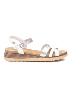 Sandalias blancas y plateadas con tiras, con una suela interior acolchada en tono marrón claro, cierre con hebilla y una suela de cuña texturizada en marrón.