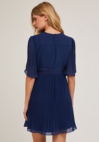 Navyblaues, plissiertes Kleid mit kurzen, transparenten Ärmeln, taillierter Passform und Knopfdetails im Rücken. Hergestellt aus leichtem Stoff mit fließender Silhouette.