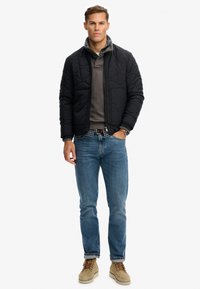 Mężczyzna w czarnej pikowanej kurtce noszonej na warstwowych koszulach, niebieskich dżinsach, beżowych zamszowych butach i zegarku na nadgarstku, stojący na tle jednolitego białego tła.