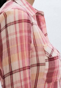 Gros plan sur une personne portant une chemise à carreaux rose, beige et rouge, avec un col et une poche poitrine.