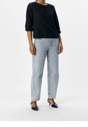 Femme portant un chemisier noir à manches longues, un jean gris clair taille haute et des escarpins noirs à bout pointu avec des brides autour de la cheville, debout sur un fond blanc.