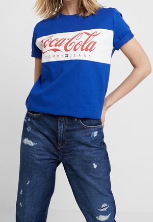 Vrouw draagt een blauw T-shirt met een rood Coca-Cola Tommy Jeans-logo en versleten donkerblauwe jeans tegen een effen achtergrond.
