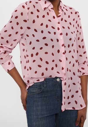 Osoba ubrana w jasnoróżową koszulę na guziki z wzorem ciemnoczerwonych ust i podwiniętymi rękawami, w połączeniu z niebieskimi dżinsami.