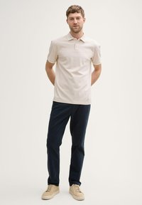 Polohemd in hellem Beige mit einem Kragen und drei Knöpfen, ergänzt durch grüne Akzente; kombiniert mit dunkelblauen Hosen und beigefarbenen Schuhen.