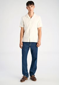 Camisa de botones de manga corta en color beige claro con cuello plano, combinada con jeans azul oscuro y zapatos marrones de cordones con suela texturizada.