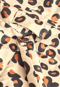 Beige tyg med ett leopardmönster som har svarta och orange former, med synliga sömmar och en metallknappdetalj.