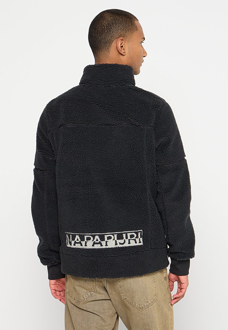 Schwarze Fleecejacke mit hohem Kragen, strukturiertem Material und sichtbarem Logodetail auf der Rückseite, kombiniert mit beigen Hosen.