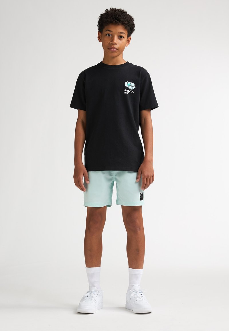 Schwarzes Baumwoll-T-Shirt mit einem Frontmotiv, kombiniert mit mintgrünen Badeshorts mit einem schwarzen Etikett. Weiße Sportschuhe und Socken.