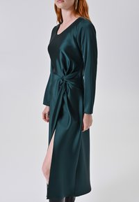 Abito lungo in satin verde smeraldo con scollatura a V, maniche lunghe e un dettaglio attorcigliato in vita; presenta uno spacco alto sulla coscia su un lato.