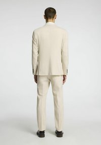Beżowy garnitur z dopasowaną marynarką i prostymi spodniami, wykonany z gładkiego materiału o subtelnej fakturze. Widok z tyłu ukazuje przesuwane szwy ramion.