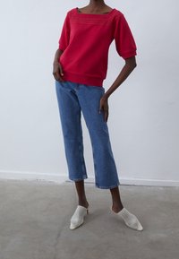 Pullover rosso con scollatura testurizzata, abbinato a jeans cropped blu e mule in rete bianca. Tessuto morbido e vestibilità rilassata.