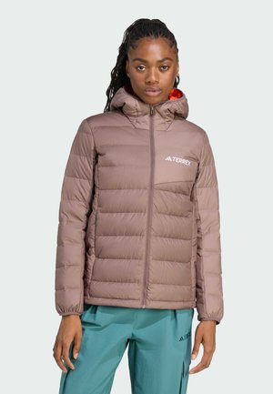 Vrouw in een mauve pufferjack met capuchon en teal broek, staande rechtop tegen een effen lichte achtergrond.