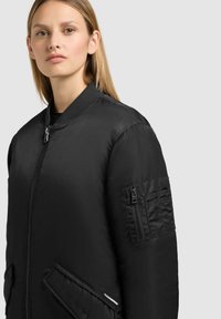 Giacca bomber nera con una finitura liscia e lucida, colletto a costine, tasche anteriori e tasca con zip sulla manica decorata con dettagli di cucitura.
