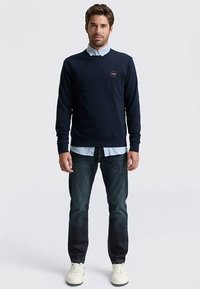 Tengerészkék kerek nyakú pulóver bordázott mandzsettákkal és szegéllyel, világoskék galléros ing fölött viselve, sötét farmernadrággal és fehér sportcipővel párosítva.