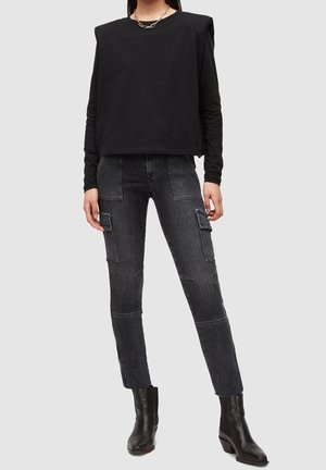 Osoba ubrana w czarną, długorękawową bluzkę z wypchanymi ramionami, ciemnoszare jeansy bojówki z przednimi kieszeniami oraz czarne botki na obcasie.