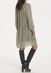 Robe à manches longues, longueur genou, en tissu beige clair avec un motif abstrait noir. Assortie à des bottes hautes en cuir noir.