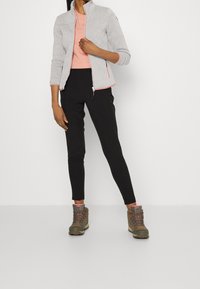 Hellgraue Fleecjacke mit Reißverschluss über einem rosa T-Shirt, kombiniert mit schwarzen Hosen und braunen Wanderschuhen mit rosa Akzenten.