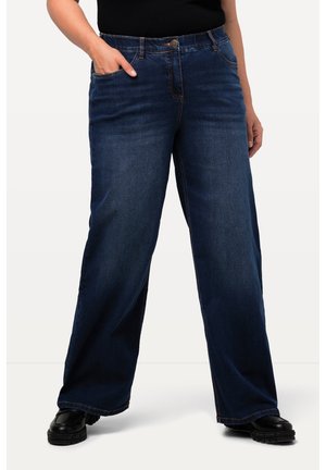 Personne portant un jean large bleu foncé, des chaussures noires épaisses et un haut noir, avec une main dans la poche, debout devant un fond blanc.