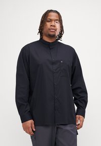 Camicia nera con colletto alla cinese, maniche lunghe, taschino sul petto e un piccolo logo sulla tasca. Texture liscia, design elegante.