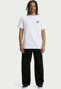 Tricou alb din bumbac cu un logo rotund negru pe partea stângă a pieptului, purtat cu pantaloni negri largi și adidași negri, stând în fața unui fundal deschis.