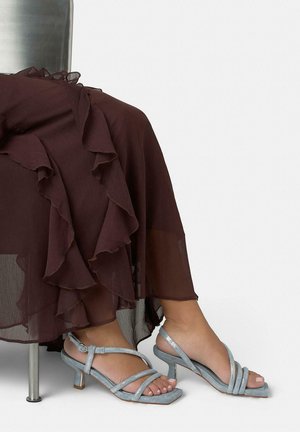 Pies con sandalias plateadas de tacón bajo y tiras, combinadas con una falda marrón semitransparente con volantes, sentados en un taburete metálico contra un fondo blanco.