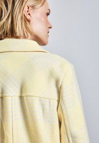 Gul vävd jacka med en texturerad yta, krage och ett subtilt geometriskt mönster i ljusgrått.