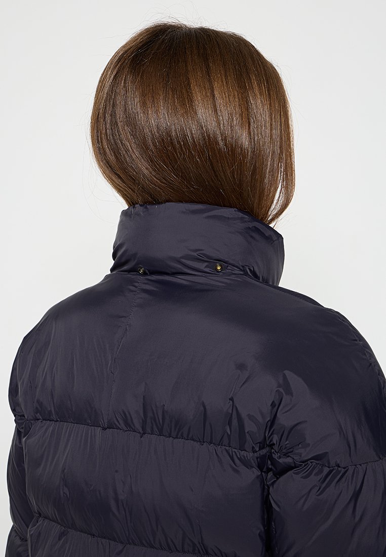 Person mit glattem braunem Haar, der sich abwendet und eine hochkragen, dunkle marineblaue Daunenjacke trägt. Der Hintergrund ist einfarbig hellgrau.
