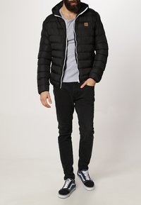 Chaqueta negra acolchada con capucha, cierre de cremallera y detalles en blanco, llevada sobre una camiseta gris, combinada con pantalones negros y zapatillas.