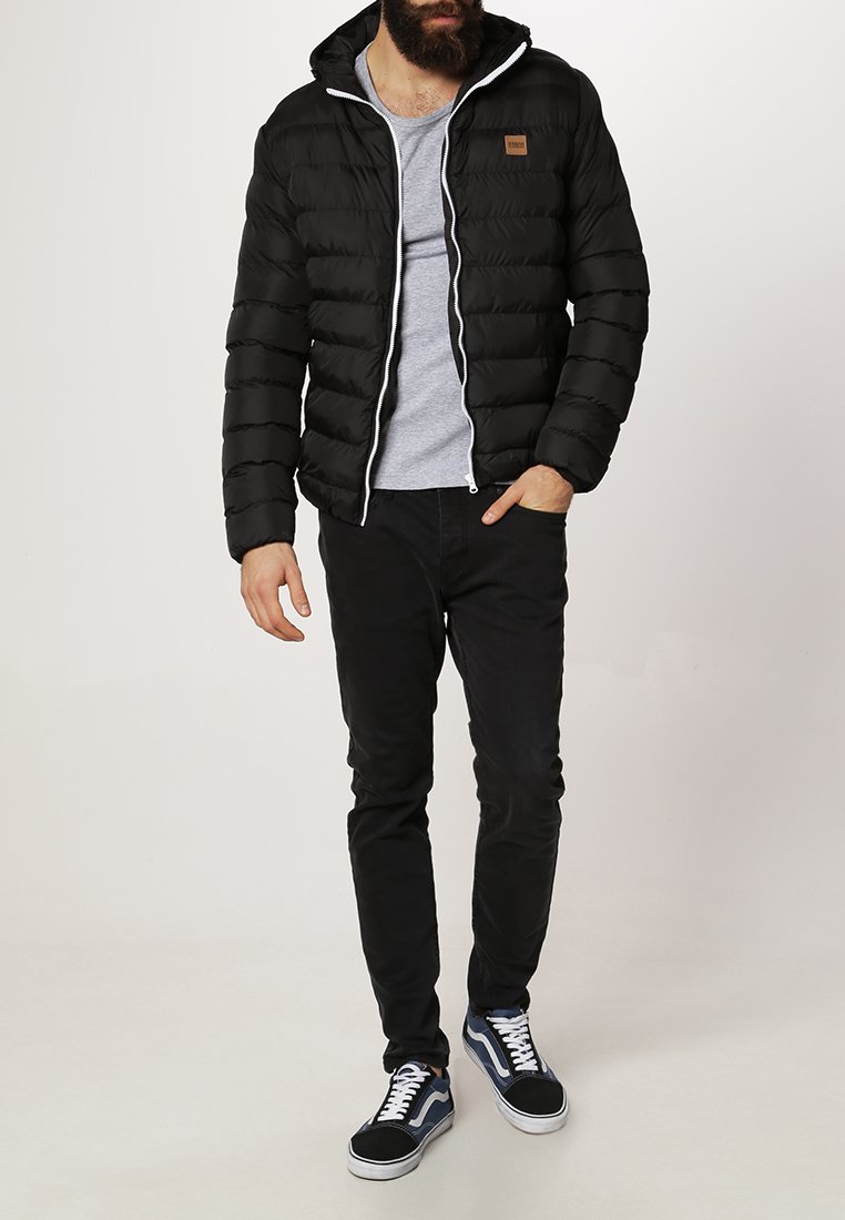 Chaqueta negra acolchada con capucha, cierre de cremallera y detalles en blanco, llevada sobre una camiseta gris, combinada con pantalones negros y zapatillas.