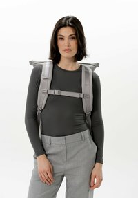 Zaino grigio con spalline regolabili e fibbie indossato sopra una maglietta a maniche lunghe aderente. Il modello ha i capelli scuri e indossa pantaloni grigi chiari.