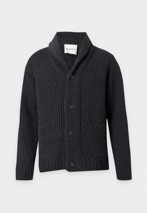 Homme portant un cardigan noir à col châle boutonné, chemise blanche, jean large bleu clair et chaussures noires, debout les mains dans les poches.